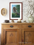 Pastel painting print of a pygmy owl on a lichen covered branch with a mahogany frame and 2” white mat hanging over a natural elements credenza.  Rendered in a realistic style by award winning artist Kathie Miller.