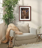 Pastel painting print of a young giraffe with a mahogany frame and 2” white mat hanging in a cozy sitting corner.  Rendered in a realistic style by award winning artist Kathie Miller.