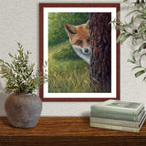Pastel portrait print of a shy fox peeking from behind a tree with a mahogany frame and 2” white mat hanging over a side board.  Rendered in a realistic style by award winning artist Kathie Miller.