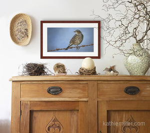 Pastel portrait print of a Yellow Rumped Warbler on a barbed wire fence with a mahogany frame and 2” white mat hanging over a natural elements credenza.  Rendered in a realistic style by award winning artist Kathie Miller.