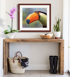 Pastel portrait print of a toucan with a mahogany frame and 2” white mat hanging in a rustic style entrance hall. Rendered in a contemporary style using bold strokes and bright colors by award winning artist Kathie Miller. 