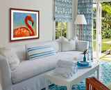 A mock up photo of a living room decorated in a beach theme with blues and whites. Hung on the wall is a print of my painting "Cora-Caribbean Flamingo" by award winning artist Kathie Miller. This is a contemporary pastel portrait of a flamingo using bright reds, orange and yellows using expressive strokes. The background is bright blue fading down to a soft blue-green.