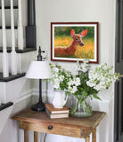 Pastel portrait print of a fawn lying in the grass in the morning sun with a simple wood frame and 2” white mat hanging in stairs nook. Rendered in a contemporary style using bold strokes and bright colors by award winning artist Kathie Miller. 