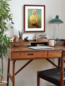 A mock up photo of a home office. Hung on the wall is a print of my painting “Daisy-Duckling” by award winning artist Kathie Miller. The print has a mahogany frame and white mat. This is a contemporary pastel portrait of a little yellow duckling rendered in bold expressive strokes and bright colors. 