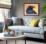 Pastel portrait print of a raven with a mahogany frame and 2” white mat hanging in a cozy sitting room. Rendered in a contemporary style using bold strokes and bright colors by award winning artist Kathie Miller. 