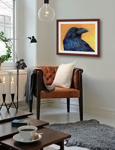 Pastel portrait print of a raven with a mahogany frame and 2” white mat hanging in a modern living room corner. Rendered in a contemporary style using bold strokes and bright colors by award winning artist Kathie Miller. 