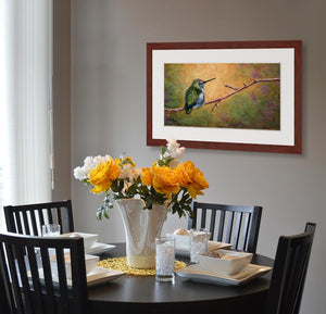 Pastel portrait print of a hummingbird resting on a branch with the morning light behind it framed in mahogany and a white mat  hanging in a casual dining room.  Rendered in a contemporary style using bold strokes and bright colors by award winning artist Kathie Miller.
