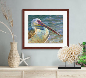 Pastel portrait of a pelican with the ocean in the background. Print with mahogany frame and a 2” white mat  hanging in a beach décor side bar.  Rendered in a contemporary style using bold strokes and bright colors by award winning artist Kathie Miller.