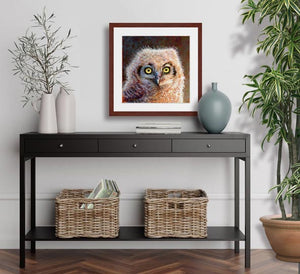 Pastel portrait print of a fluffy great horned owl chick with a mahogany frame and 2” white mat hanging in an entrance hall. Rendered in a contemporary style using bold strokes and bright colors by award winning artist Kathie Miller. 