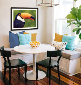 Pastel portrait print of a toucan with a simple dark wood frame and 2” white mat hanging in sunny breakfast nook. Rendered in a contemporary style using bold strokes and bright colors by award winning artist Kathie Miller. 