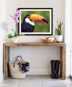 Pastel portrait print of a toucan with a simple dark wood frame and 2” white mat hanging in an entrance hall. Rendered in a contemporary style using bold strokes and bright colors by award winning artist Kathie Miller. 