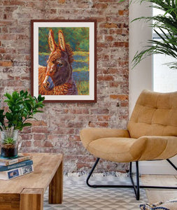 Pastel portrait print of  a donkey in the sun with a mahogany frame and 2” white mat hanging in a living room corner sitting area. Rendered in a contemporary style using bold strokes and bright colors by award winning artist Kathie Miller. 